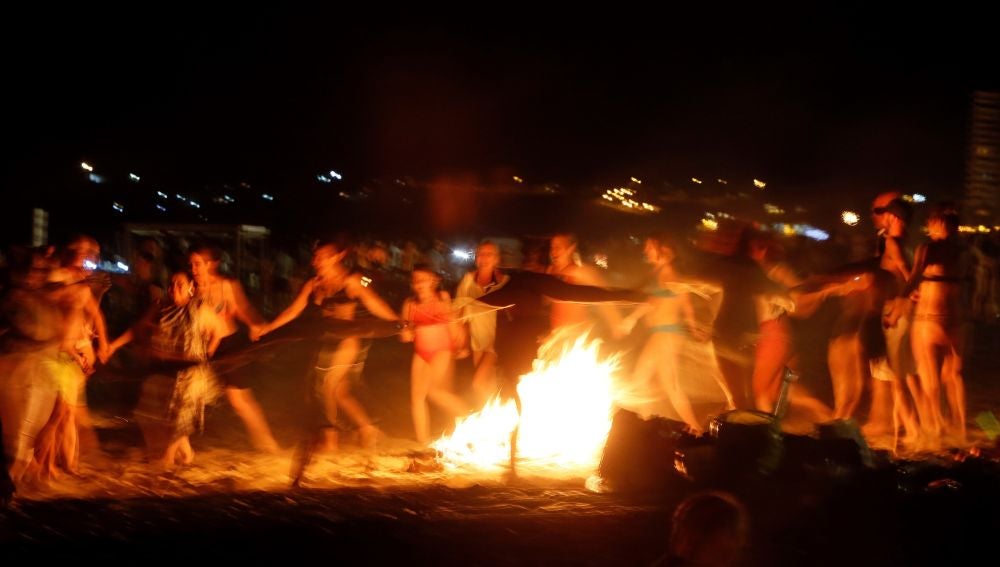 Un grupo de personas baila alrededor de una hoguera en la playa.