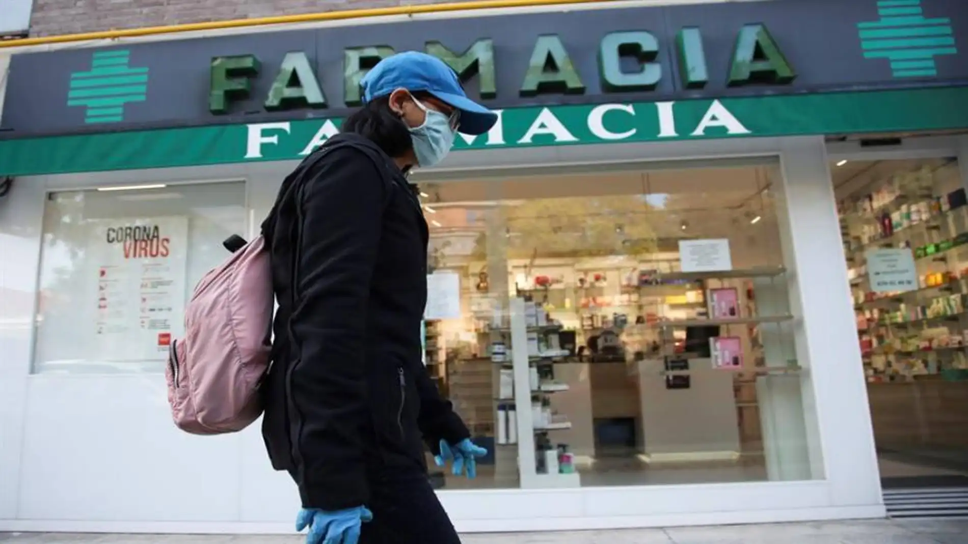 Una viandante, protegida con guantes y mascarilla, pasa por delante de una farmacia.