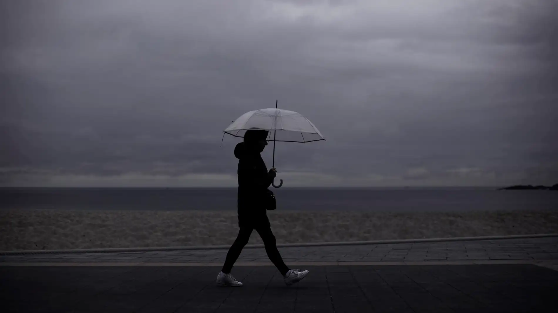 Una mujer camina por una playa mientras llueve. / Efe