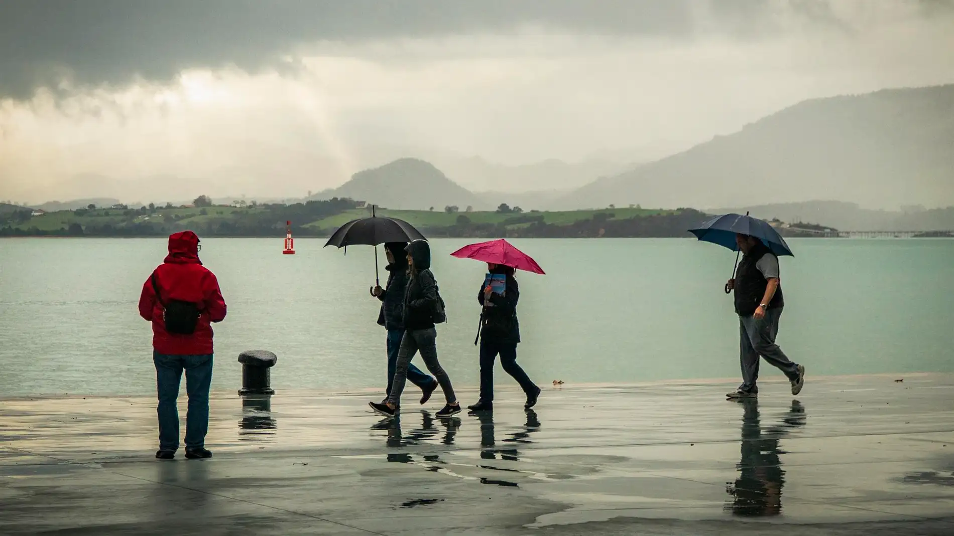 Gente pasea bajo la lluvia junto a la bahía de Santander.