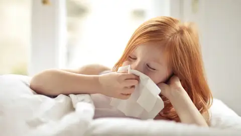 Niña pelirroja descansando en la cama, limpiándose la nariz con un tissue, indispuesta con síntomas de gripe Niña pelirroja descansando en la cama, limpiándose la nariz con un tissue, indispuesta con síntomas de gripe