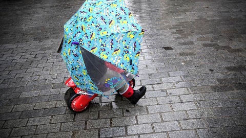 Un ni&ntilde;o se protege de la lluvia bajo un paraguas mientras disfruta con su moto de juguete en una calle de Pamplona 