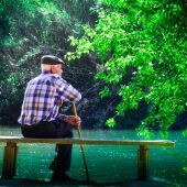 Fotografía de un hombre de avanzada edad, con bastón y boina, sentado en un banco frente a un estanque, en un parque.