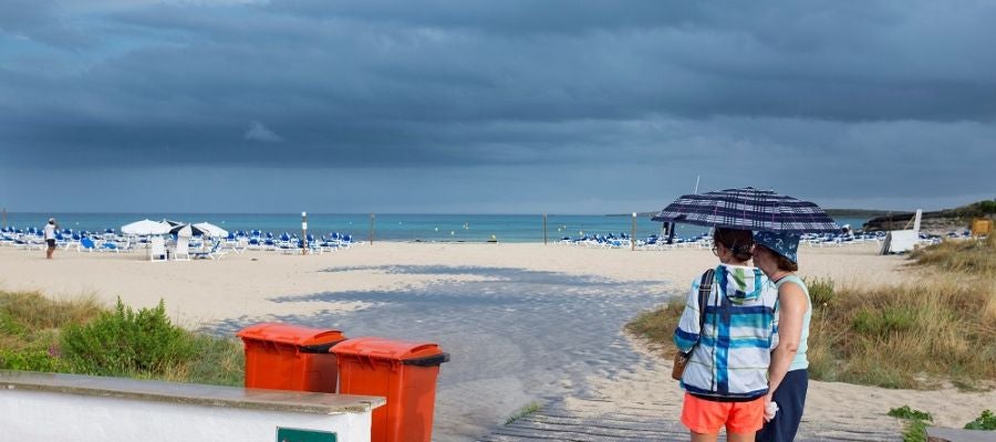 Dos mujeres se resguardan de la lluvia bajo un paraguas en una playa de Menorca Dos mujeres se resguardan de la lluvia bajo un paraguas en una playa de Menorca