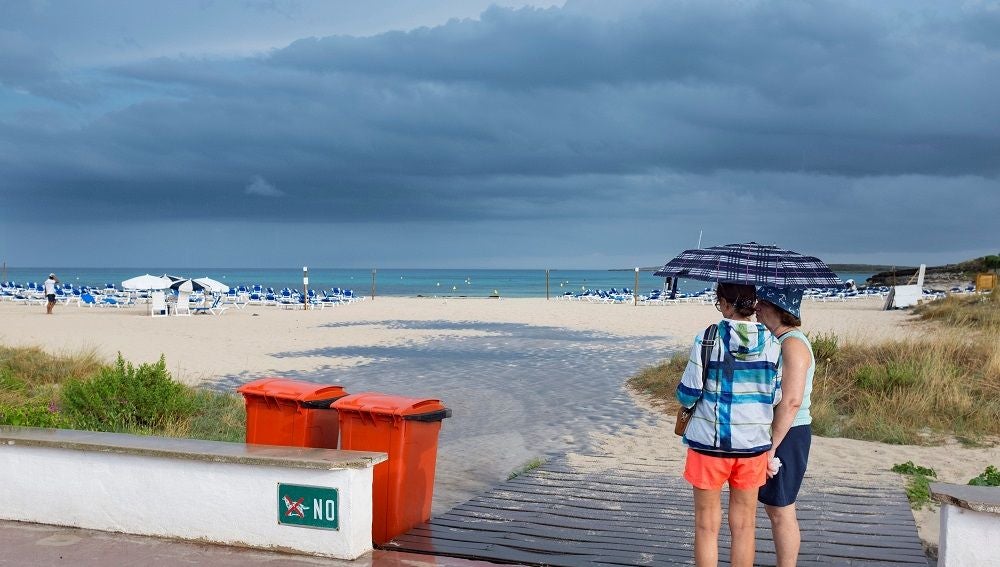 Dos mujeres se resguardan de la lluvia bajo un paraguas en una playa de Menorca