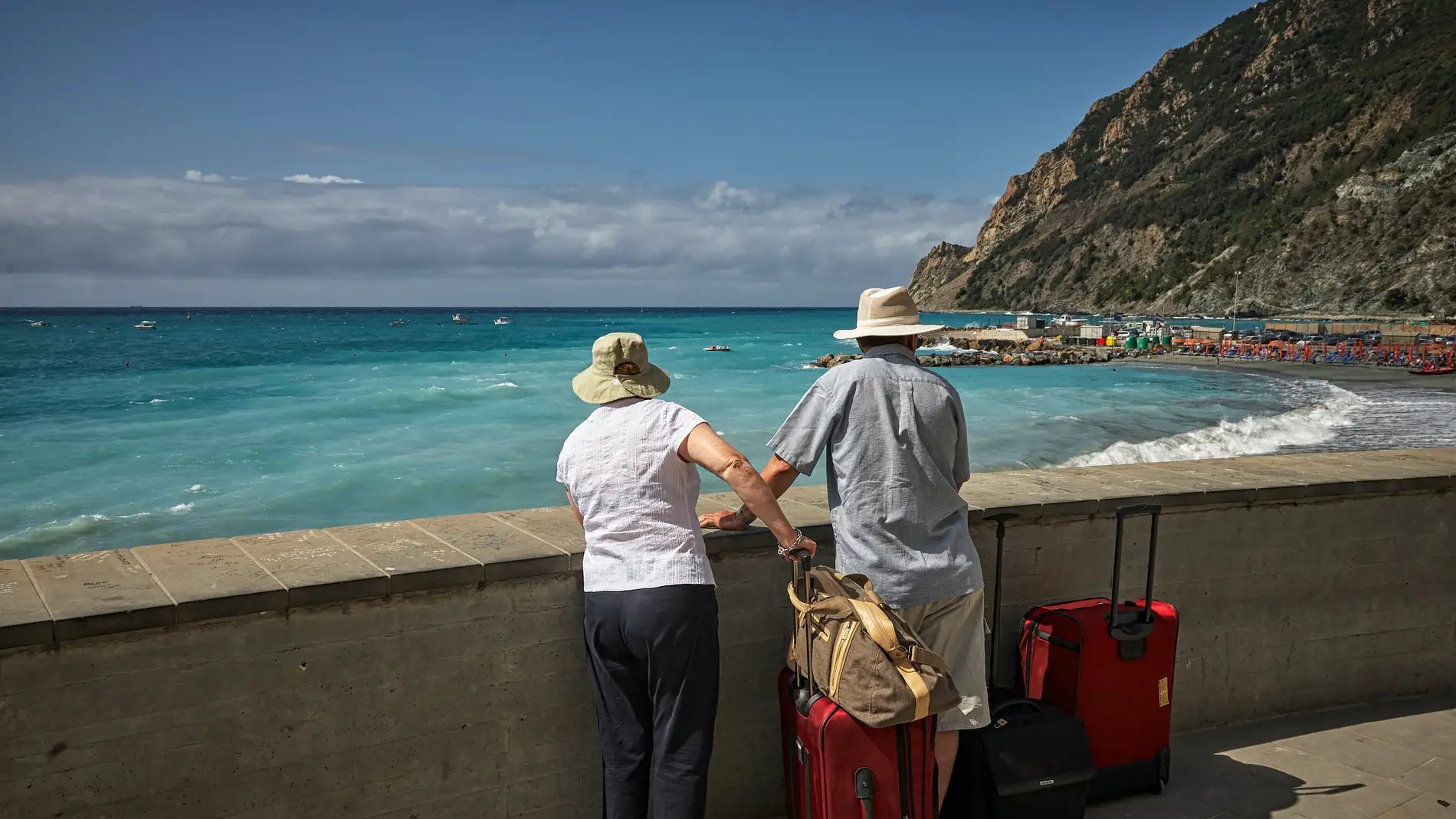 Imagen de archivo de dos personas mayores, de espaldas, mirando al mar junto a un par de maletas rojas/ Unsplash