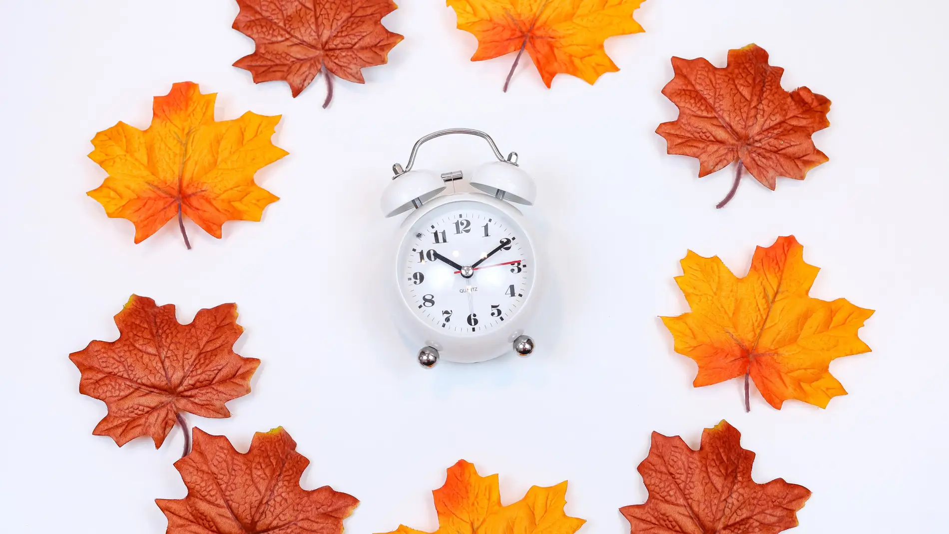 Imagen de archivo de un reloj blanco rodeado de hojas con colores otoñales/ Unsplash