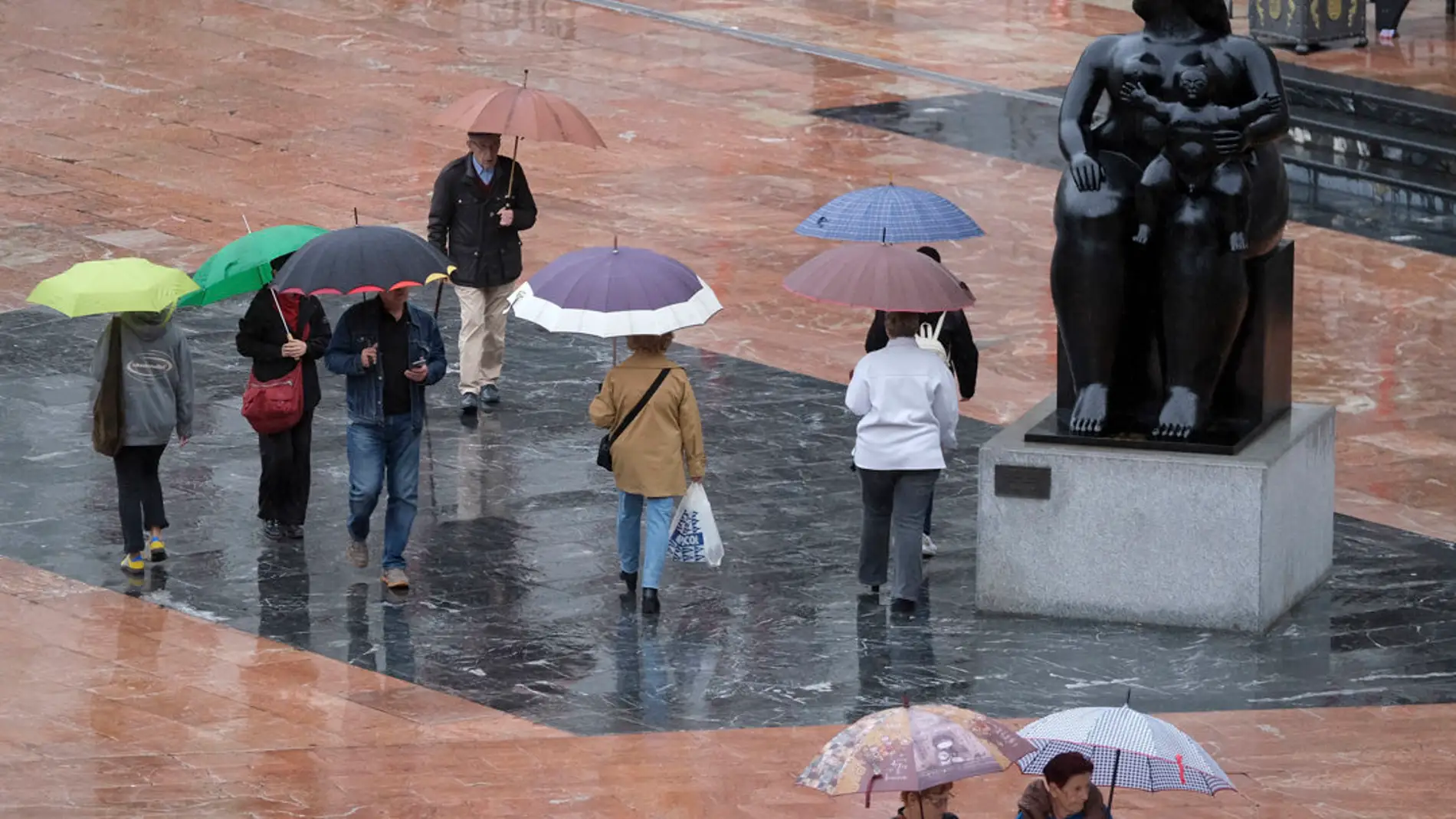 Viandantes con paraguas bajo la lluvia junto a La Maternidad de Botero. EFE/Paco Paredes