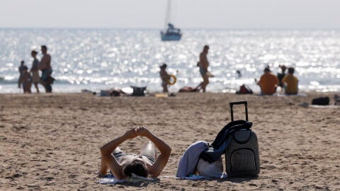 Imagen de archivo de un turista con su maleta tomando el sol en una playa valenciana este verano. 