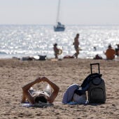 Imagen de archivo de un turista con su maleta tomando el sol en una playa valenciana este verano. 