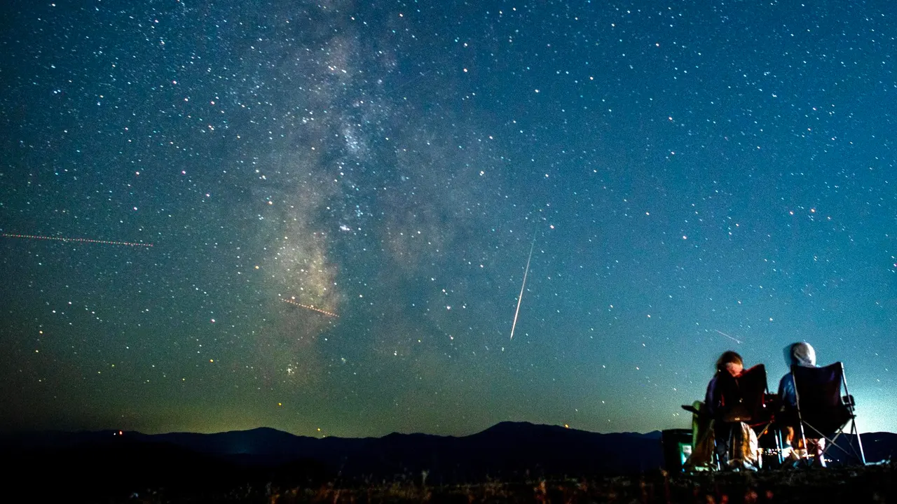 Una pareja observa cómo un meteoro surca el cielo nocturno más allá de la Vía Láctea durante las Perseidas.
