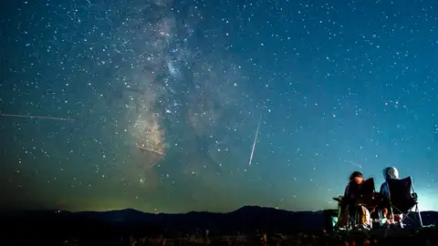 Una pareja observa cómo un meteoro surca el cielo nocturno más allá de la Vía Láctea durante las Perseidas. Una pareja observa cómo un meteoro surca el cielo nocturno más allá de la Vía Láctea durante las Perseidas.