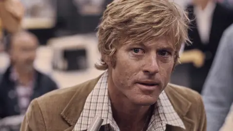 El actor estadounidense Robert Redford, con un blazer de pana marrón sobre una camisa a cuadros, recrea las oficinas de The Washington Post en un fotograma publicitario de "Todos los hombres del presidente", filmado en los estudios Burbank de Burbank, California, en 1976. El actor estadounidense Robert Redford, con un blazer de pana marrón sobre una camisa a cuadros, recrea las oficinas de The Washington Post en un fotograma publicitario de "Todos los hombres del presidente", filmado en los estudios Burbank de Burbank, California, en 1976.